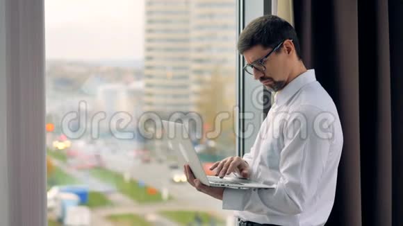 忧郁的商人在窗户附近使用笔记本电脑视频的预览图