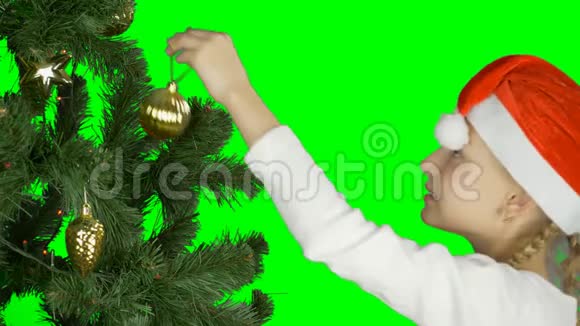 戴着红色圣诞帽的年轻女孩用金色球体装饰绿色圣诞树准备庆祝一下视频的预览图
