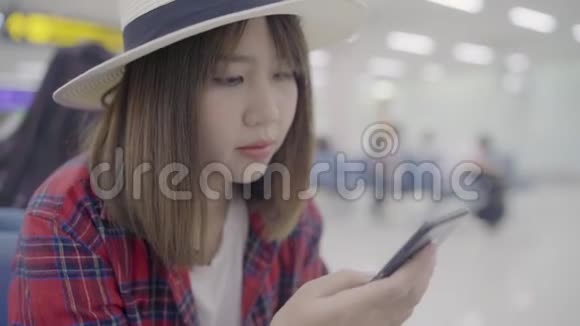 快乐的亚洲女人在机场等待航班时坐在候机大厅的椅子上使用和检查她的智能手机视频的预览图