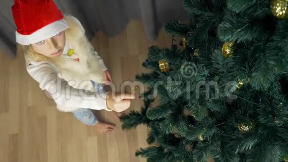 快乐的年轻女孩戴着红色圣诞老人帽子有趣而快乐装饰圣诞树的白人女孩金球落到地板附近视频的预览图