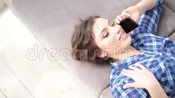 迷人的年轻女子躺在沙发上打着电话年轻女子打着手机微笑着视频的预览图