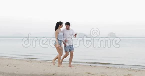 一对夫妇在海滩上用手机散步拥抱交谈年轻男女游客在网上交流视频的预览图