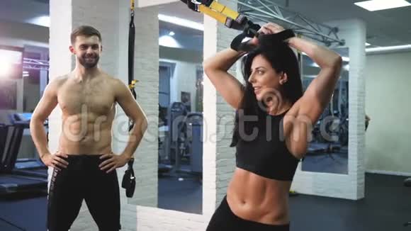 年轻有魅力的女人在健身房健身室里用健身带做基础腹肌斜向锻炼一个看运动的男人视频的预览图