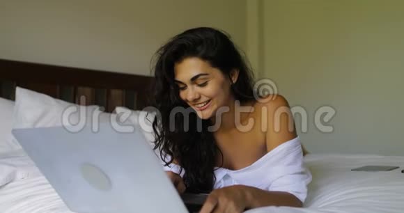 年轻的女人用笔记本电脑快乐地笑着美丽的女孩躺在卧室的早晨视频的预览图
