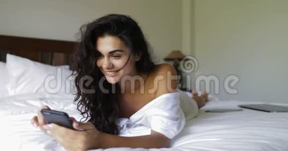 年轻的女人用手机快乐的微笑美丽的女孩躺在早晨视频的预览图