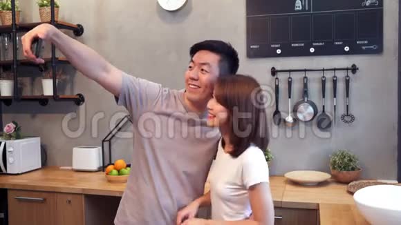 快乐的亚洲年轻夫妇在家厨房做饭时用智能手机自拍男人和女人准备健康的食物视频的预览图