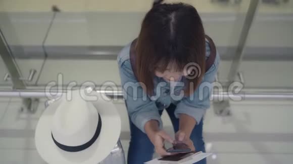 快乐的亚洲女人在机场等待航班时坐在候机大厅的椅子上使用和检查她的智能手机视频的预览图