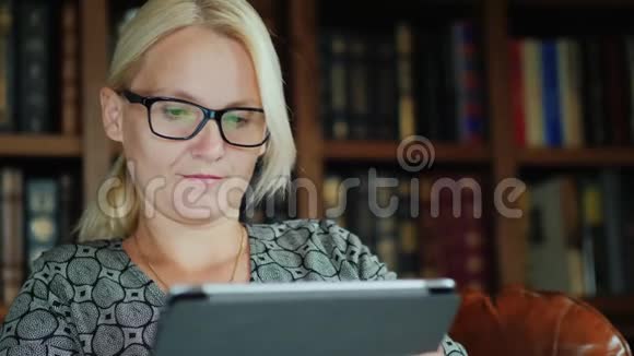 戴眼镜坐在椅子上用平板电脑的女人的肖像在书架的背景下视频的预览图