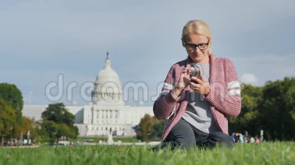 一个戴眼镜的年轻女人坐在草坪上享受着智能手机在华盛顿国会大厦的背景下视频的预览图