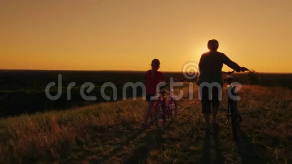 一位6岁的老太太和小女孩一起在风景如画的地方散步骑自行车日落时的后视图视频的预览图