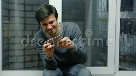男人坐在窗台上玩手机游戏视频的预览图