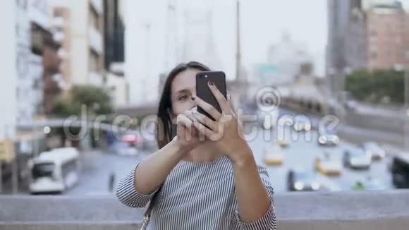年轻漂亮的女人在智能手机上自拍照片交通道路和繁忙的城市景观背景视频的预览图
