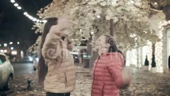 两个年轻女孩在秋天的傍晚沿着街道跳舞两个年轻女孩用耳机听音乐视频的预览图