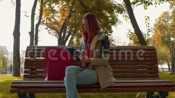 在阳光明媚的天气里长着狐毛的女孩坐在长凳上使用平板电脑并在网上订购视频的预览图