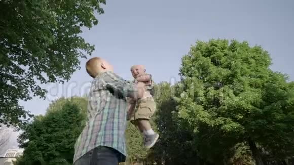 年轻的父亲牵着他的孩子的手在一个阳光明媚的日子里在公园里慢动作地把他扔到草地上视频的预览图