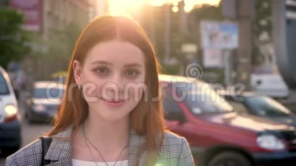 年轻的美女一头姜黄色的头发站在街上在日落时看着镜头微笑着车水马龙视频的预览图