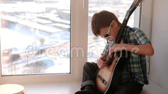 演奏乐器戴太阳镜的男孩学弹吉他坐在窗台上视频的预览图