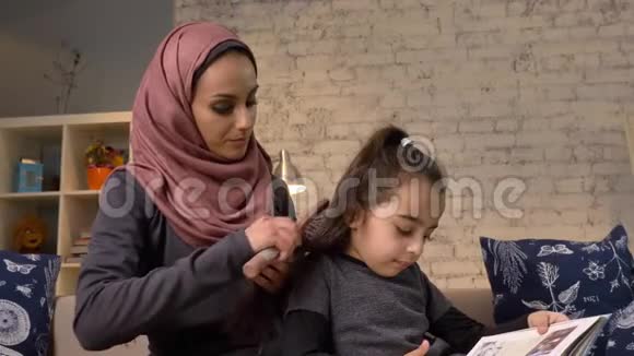 一位年轻的印度母亲戴着头巾一边梳头一边给小女儿看书一边坐在沙发上视频的预览图