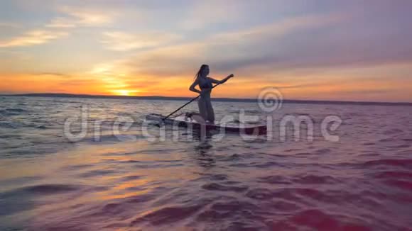 冲浪女人在冲浪板上在夕阳下漂浮在水面上视频的预览图