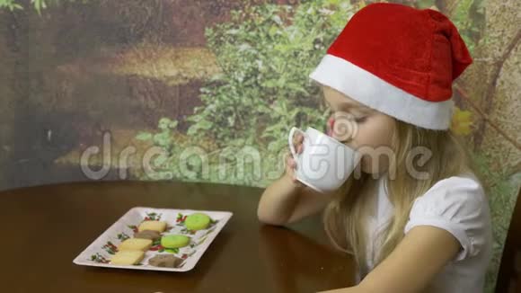 戴着红帽子的白种人年轻女孩坐在家庭厨房的桌子上女孩喝水或茶从白色杯子关门视频的预览图
