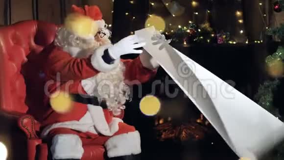 圣诞老人坐在圣诞树旁的椅子上看新年礼物单视频的预览图