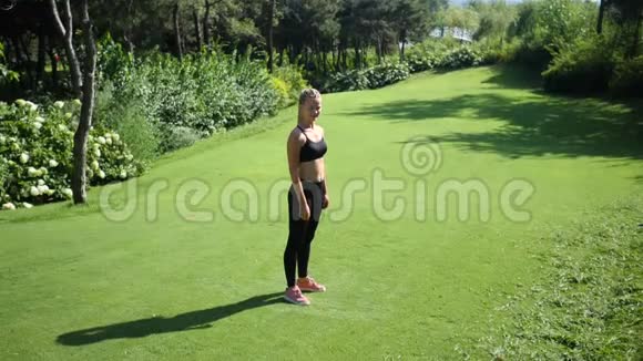 年轻的金发女人带着辫子在绿色的夏季公园的草地上做跳跃千斤顶健身锻炼慢慢视频的预览图