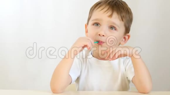 快乐的孩子微笑着在棍子上舔棒棒糖坐在白色背景的桌子旁视频的预览图