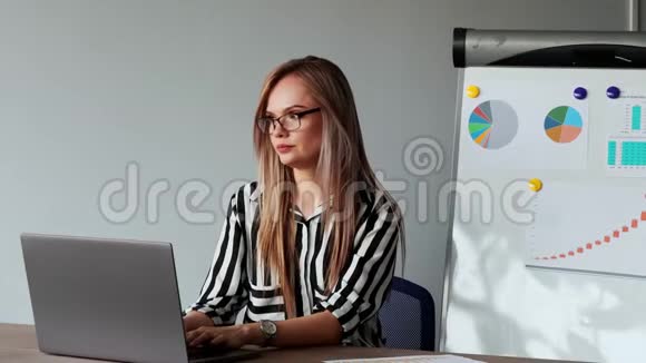 一幅美丽的欧洲女孩坐在办公室的笔记本电脑前的肖像上面有一件白色衬衫上面有图表和图片视频的预览图