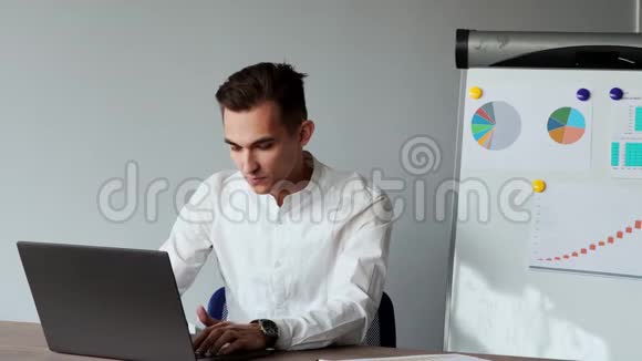 一名欧洲男性坐在办公室的笔记本电脑前在图表和桌子的背景上穿着一件白色衬衫的肖像视频的预览图