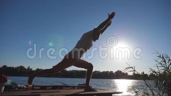 站在瑜伽馆前的运动男子剪影在湖边的一个木制码头上摆姿势在大自然训练的年轻人运动员视频的预览图