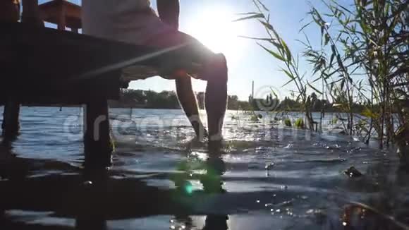 不可辨认的人坐在湖边的一个木制码头上双脚在水里摆动年轻人很放松视频的预览图