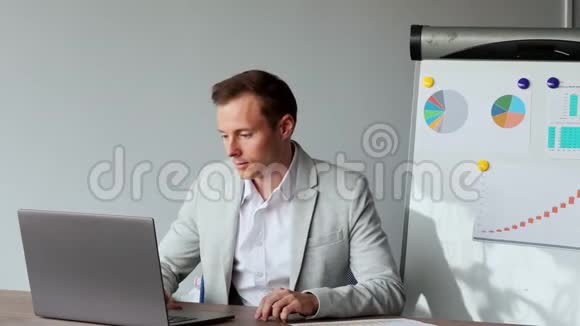 一名欧洲男性坐在办公室的笔记本电脑前在图表和桌子的背景上穿着一件白色衬衫的肖像视频的预览图