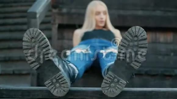 年轻的金发女孩躺在木凳上双脚朝前进入相机穿着黑色的大靴子玩她的脚放松放松视频的预览图