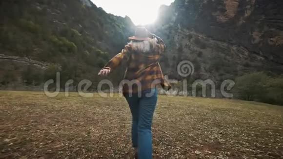 美丽的年轻女子旅行者在大自然中自由地穿越田野在群山把帽子扔到空中视频的预览图