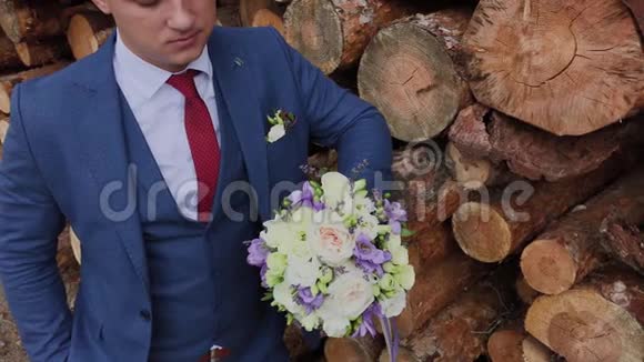 英俊的新郎穿着夹克带着一束花站在一堆原木附近视频的预览图