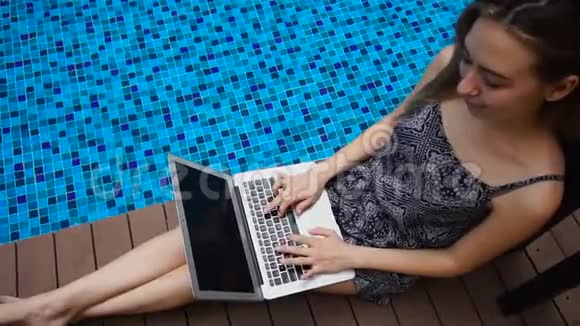 年轻女子躺在蓝色游泳池附近慢动作使用笔记本电脑视频的预览图