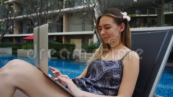 年轻女子躺在沙滩椅上在游泳池附近使用笔记本电脑视频的预览图