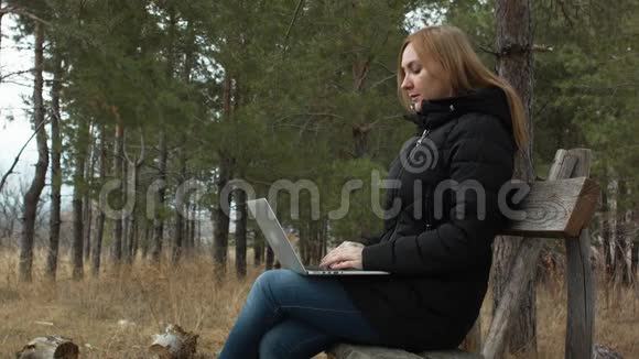 有笔记本电脑的女孩坐在森林里的公园长椅上背景是树木在露天工作秋季森林视频的预览图