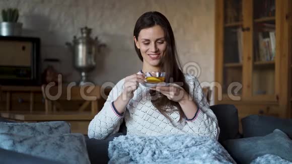 美丽的女人坐在沙发上裹着毯子喝茶视频的预览图