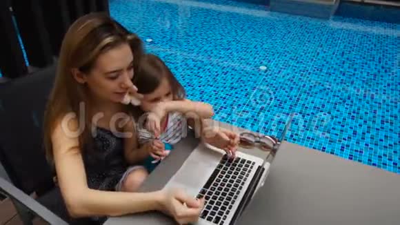 年轻的母亲和小女儿坐在游泳池附近慢动作地使用笔记本电脑视频的预览图