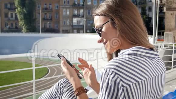 在阳光明媚的一天时尚的年轻女子坐在体育场上使用手机拍摄的特写镜头视频的预览图