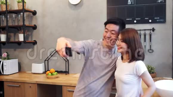 快乐的亚洲年轻夫妇在家厨房做饭时用智能手机自拍男人和女人准备健康的食物视频的预览图