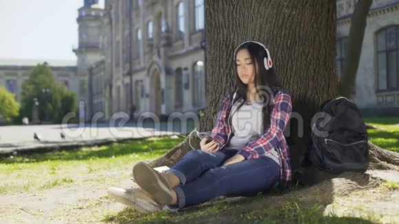 多种族女孩戴着耳机坐在树下用电话选择歌曲视频的预览图
