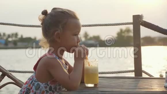 有趣的小女孩坐在河边咖啡馆的一张桌子上喝果汁视频的预览图