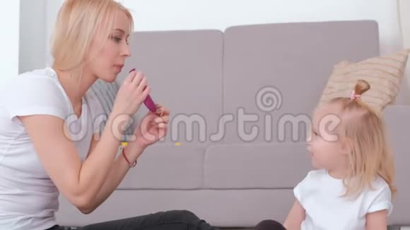迷人的年轻金发妈妈为坐在沙发附近的迷人女儿充气气球家庭消遣视频的预览图