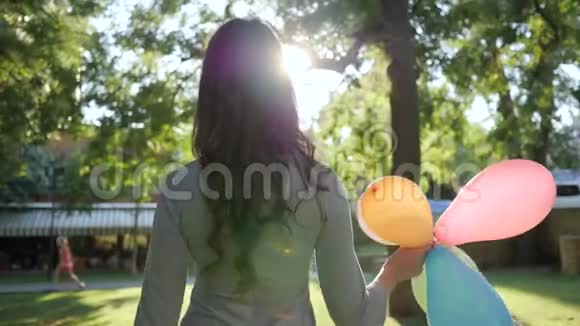 漂亮的深色头发手里拿着气球在阳光和绿树的背景下行走视频的预览图