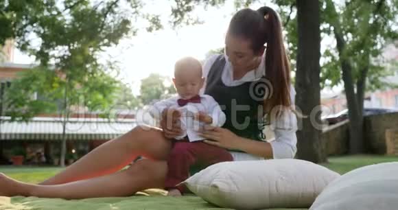 小绅士和妈妈一起坐在绿色的草坪上在公园里露天休息视频的预览图