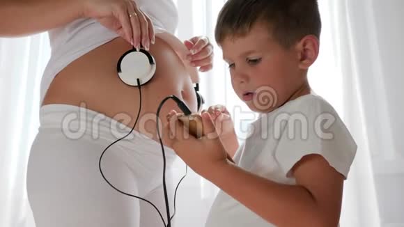 手里拿着手机的小男孩在灯火通明的房间里给孕妇戴上耳机视频的预览图