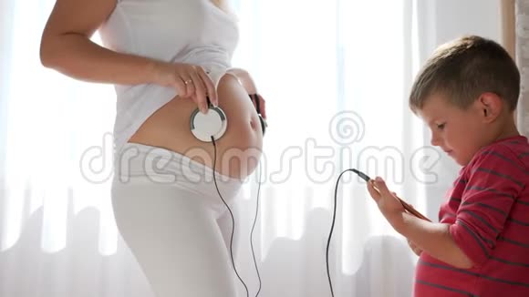 手里拿着手机的小男孩站在怀孕的女人手里把耳机放在肚子里视频的预览图
