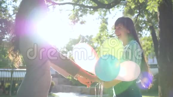 背光下的情绪快乐的女友们手牵着手在公园的背景树上旋转着五颜六色的气球视频的预览图